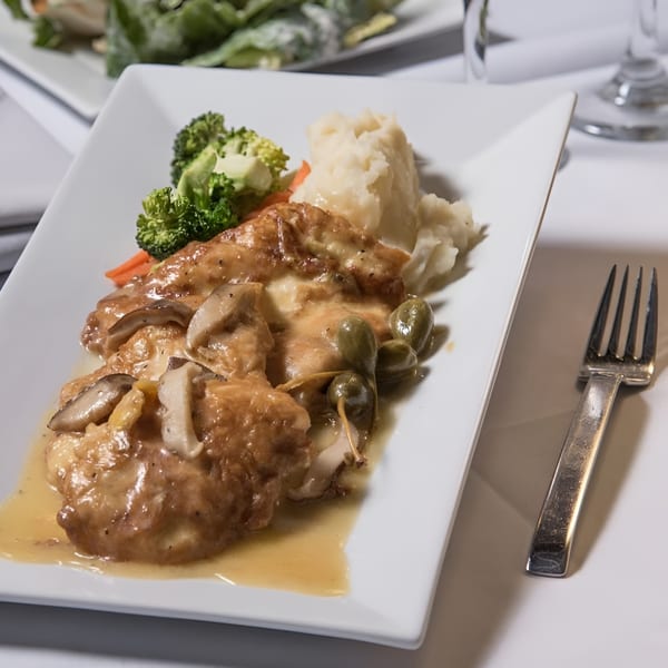 Chicken piccata with mushrooms, capers, mashed potatoes, broccoli, and carrots on a white plate.