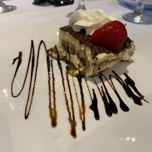 Tiramisu dessert with whipped cream and a strawberry, drizzled with chocolate sauce on a white plate.