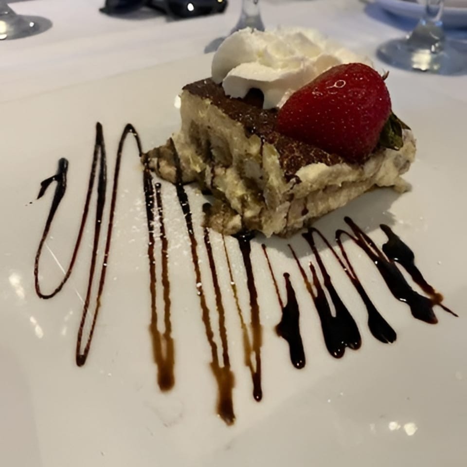 Tiramisu dessert with whipped cream and a strawberry, drizzled with chocolate sauce on a white plate.
